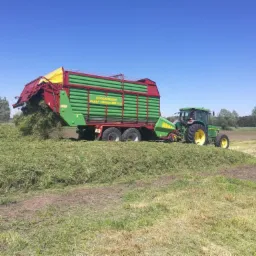 Zielony traktor John Deere z przyczepą Strautmann Super Vitesse 350+00 zbiera świeżo skoszoną trawę na polu w słoneczny dzień.