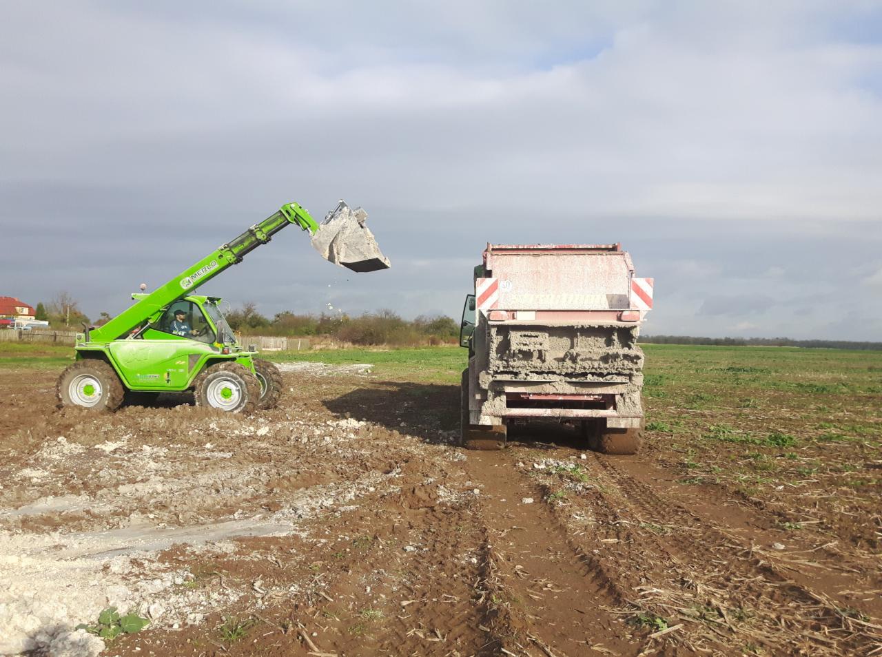 Zielona ładowarka teleskopowa Merlo z łyżką nabiera wapno i wsypuje je do brudnej, czerwono-białej posypywarki do wapna na polu w pochmurny dzień.