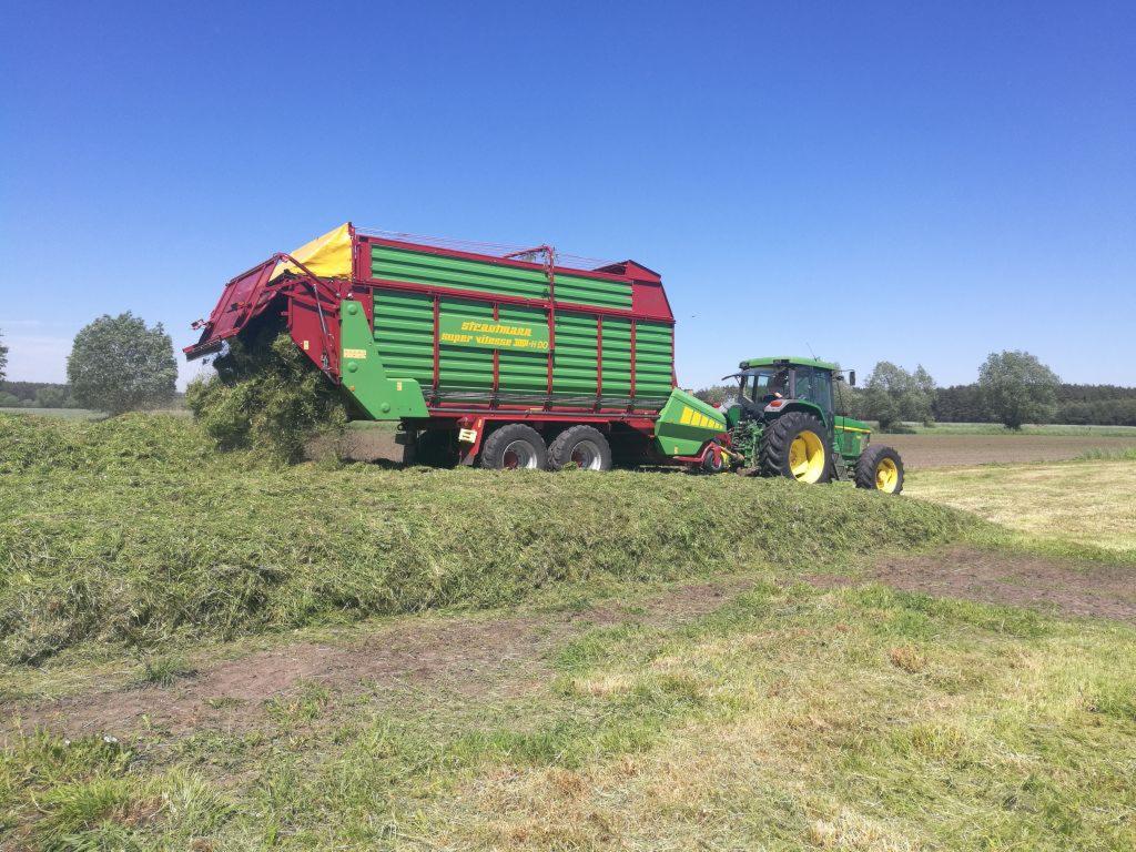 Zielony traktor John Deere z przyczepą Strautmann Super Vitesse 350+00 zbiera świeżo skoszoną trawę na polu w słoneczny dzień.