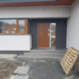 Nowoczesny dom jednorodzinny z brązowymi drzwiami wejściowymi i drewnianym oknem, widoczny fragment brukowanej ścieżki i paleta drewniana oparta o ścianę.