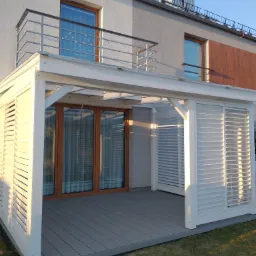 Nowoczesny taras z szarej deski kompozytowej, zabudowany białą pergolą z żaluzjowymi panelami, przylegający do budynku z balkonem.