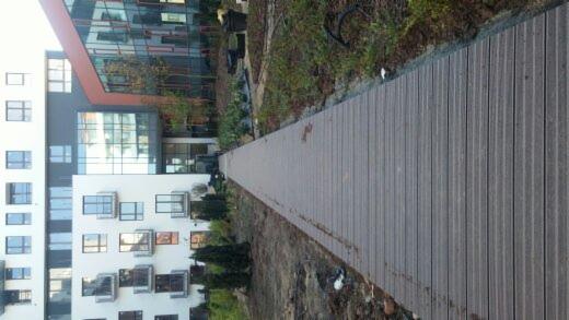 Długa, szara ścieżka z desek kompozytowych prowadząca wzdłuż budynku z licznymi oknami i zielenią.