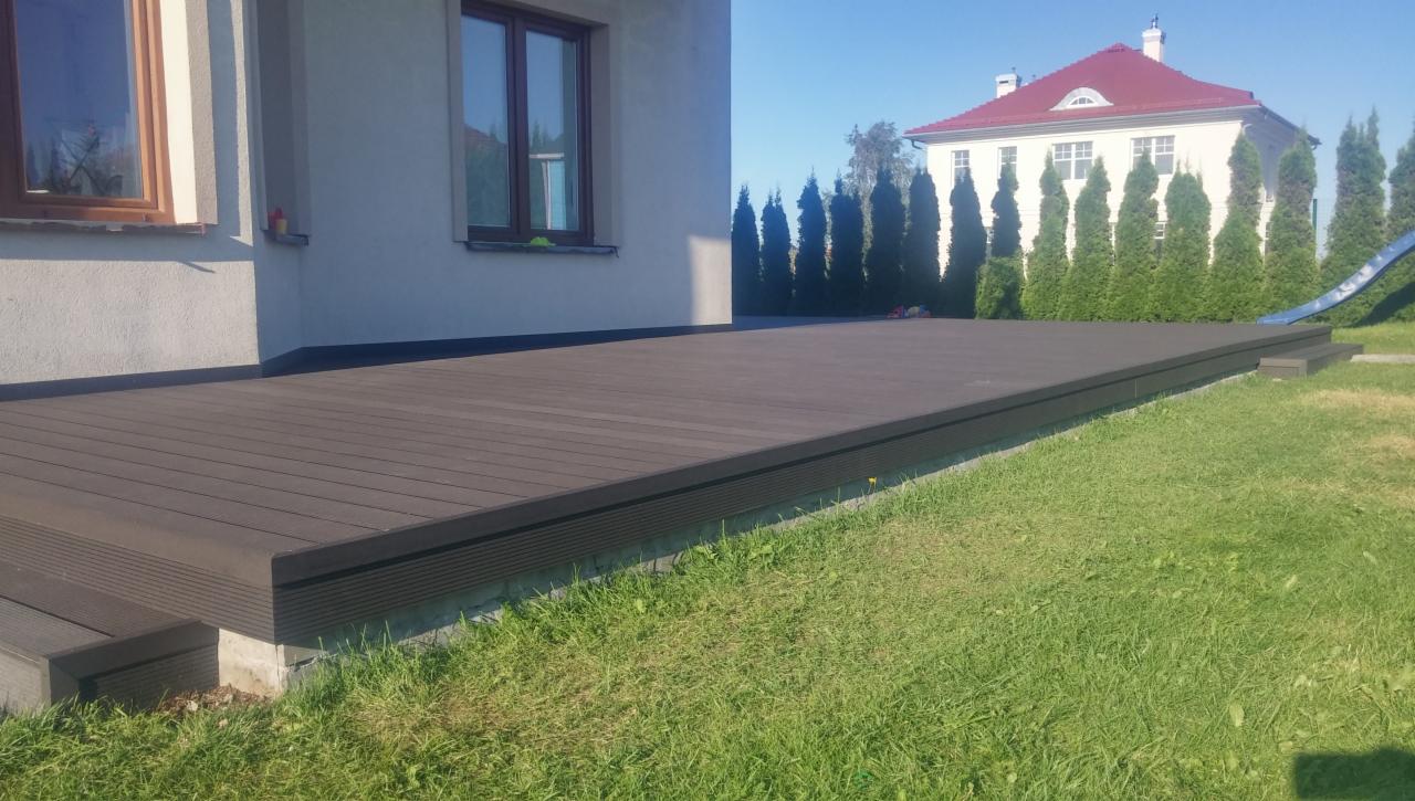 Nowoczesny taras z desek kompozytowych w kolorze ciemnego brązu, przylegający do jasnego budynku mieszkalnego z oknami, widok na zadbany trawnik i dom z czerwonym dachem w tle.