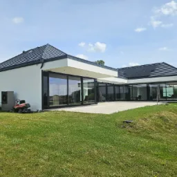 Nowoczesny dom jednorodzinny z płaskim dachem i dużymi przeszkleniami, widok z ogrodu, z boku widoczna pompa ciepła i kosiarka.