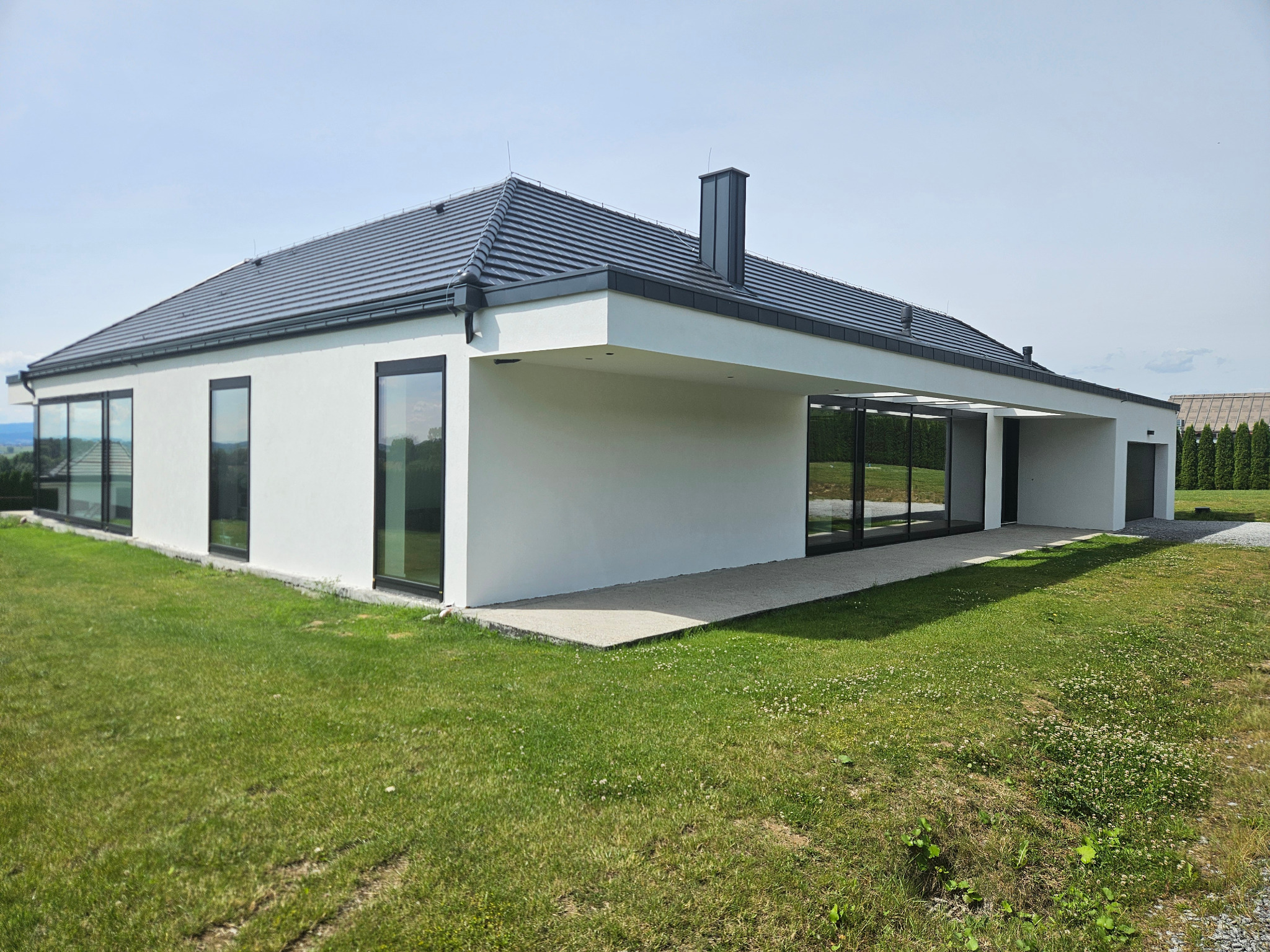 Nowoczesny, parterowy dom z płaskim dachem, białymi ścianami i dużymi oknami, otoczony zielonym trawnikiem.