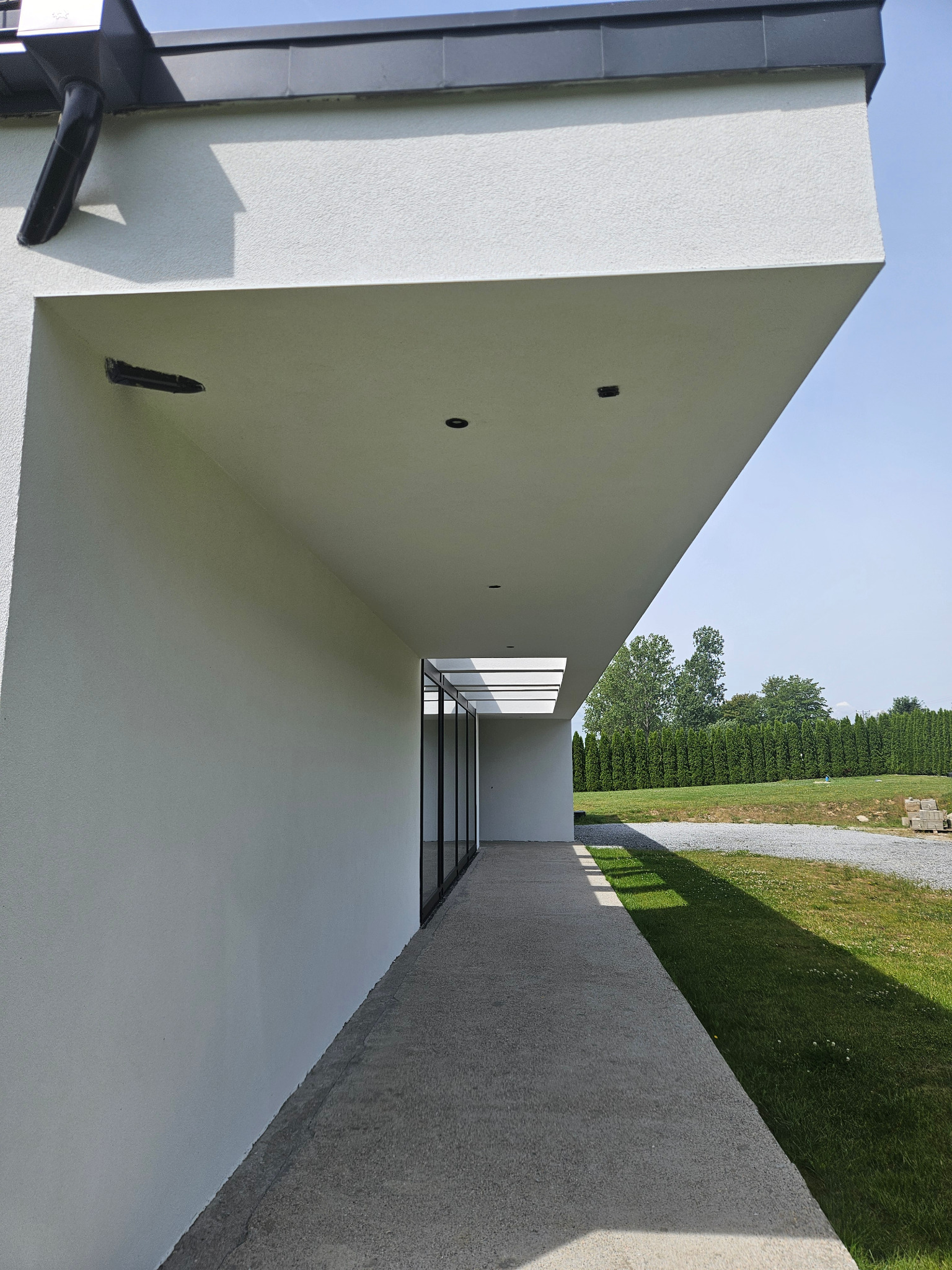 Nowoczesna elewacja domu jednorodzinnego z białym tynkiem strukturalnym, widok z boku na zadaszone przejście i starannie wypielęgnowany trawnik z tujami w tle.