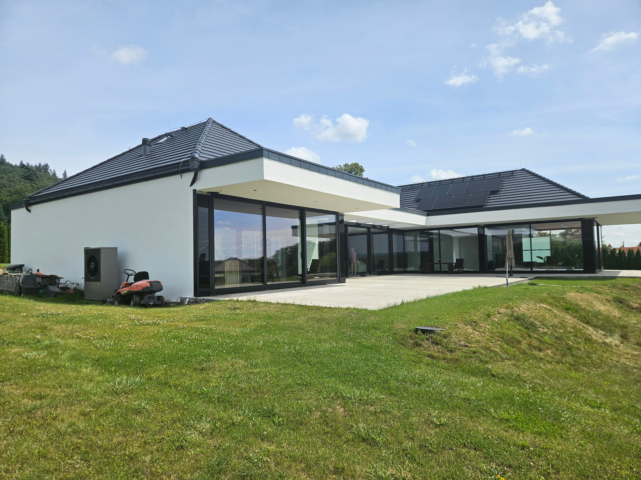Nowoczesny dom jednorodzinny z płaskim dachem i dużymi przeszkleniami, widok z ogrodu, z boku widoczna pompa ciepła i kosiarka.
