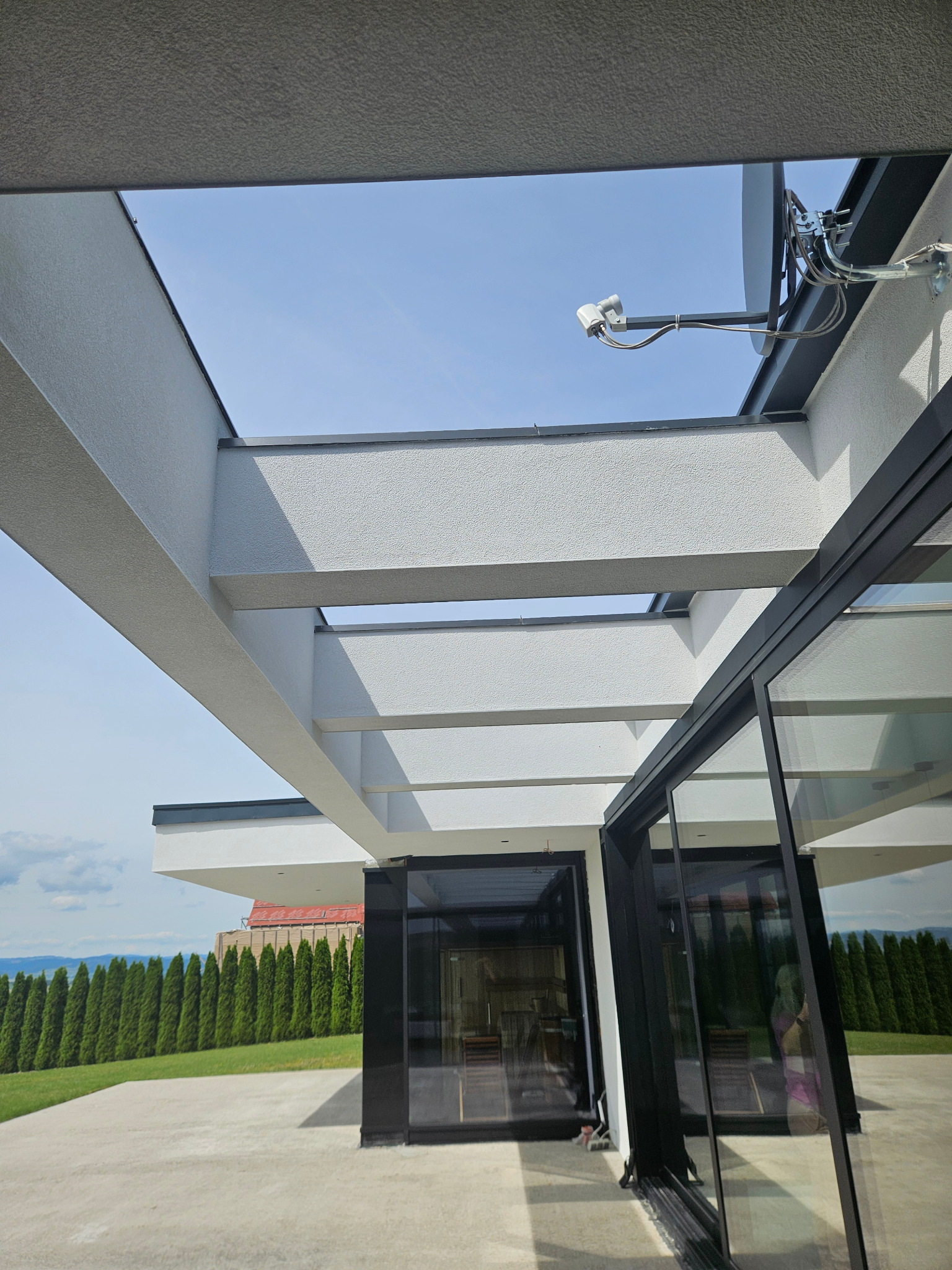 Nowoczesny dom z elewacją o jasnej, ziarnistej fakturze, widok od strony tarasu, z widoczną konstrukcją zadaszenia, czarnymi ramami okien i satelitą na ścianie.