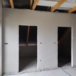 Świeżo położony, jasnoszary tynk na ścianie poddasza z widocznymi otworami na kable elektryczne i dwoma otworami drzwiowymi, surowy stan wykończenia z widoczną konstrukcją dachu i przewodami...