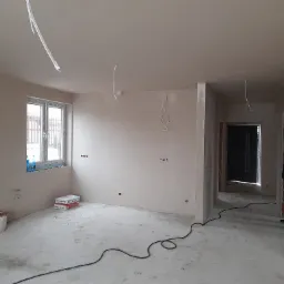 Wnętrze w stanie deweloperskim po nałożeniu gładzi, widoczne puszki elektryczne, kable zwisające z sufitu, wiadro i worki z gładzią Baulit przy oknie, surowy beton na podłodze.