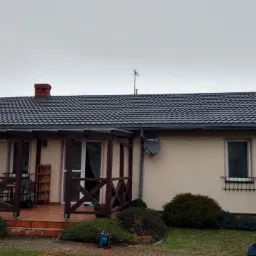 Dom jednorodzinny z nowym, ciemnoszarym dachem z blachodachówki, widoczny komin z czerwonej cegły, antena satelitarna i pergola z drewnianą konstrukcją na tarasie.