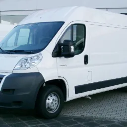 Biały bus Citroen Jumper zaparkowany na kostce brukowej, widoczny z przodu pod lekkim kątem, z czarnymi elementami nadwozia i logo marki.