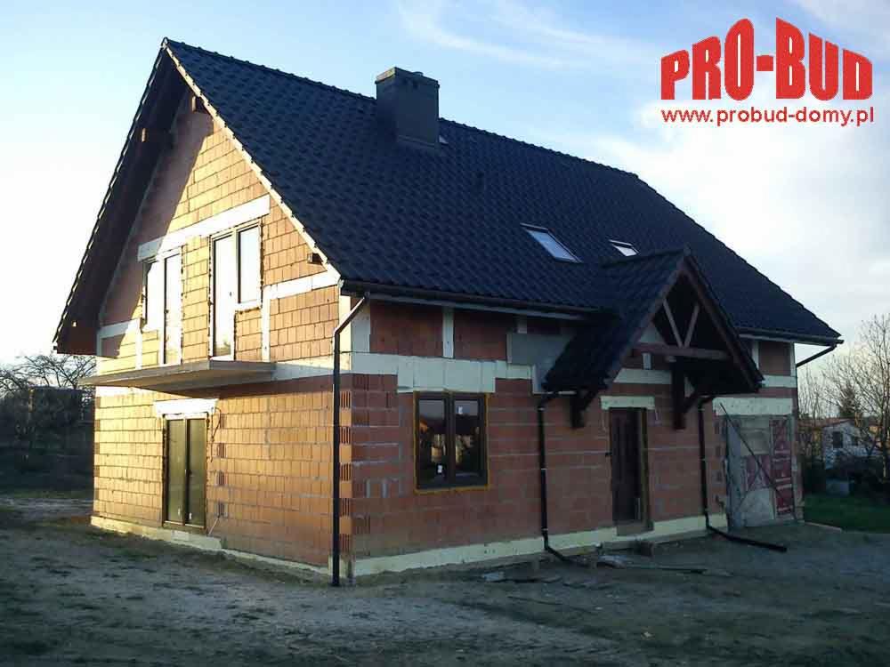Surowy budynek dwukondygnacyjny z ciemną dachówką, widoczną strukturą cegieł i oknami. Na dachu okna połaciowe. W tle drzewa i logo firmy PRO-BUD.