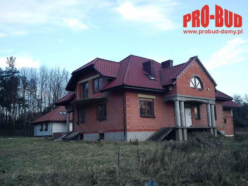 Niedokończony dom z czerwonej cegły z kolumnami wejściowymi i czerwoną dachówką, w otoczeniu trawy i drzew. Widoczne logo firmy budowlanej w prawym górnym rogu.