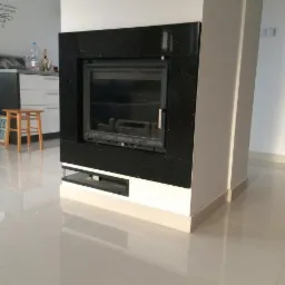 Czarny kominek z obudową z kamienia kominkowego, widoczny w salonie z błyszczącą podłogą i fragmentem kuchni w tle.