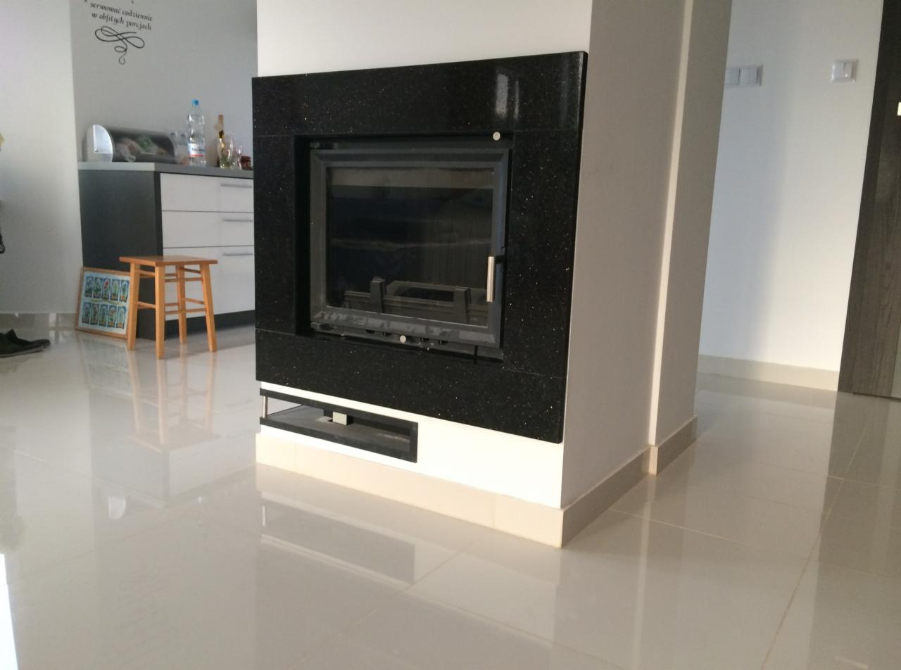 Czarny kominek z obudową z kamienia kominkowego, widoczny w salonie z błyszczącą podłogą i fragmentem kuchni w tle.