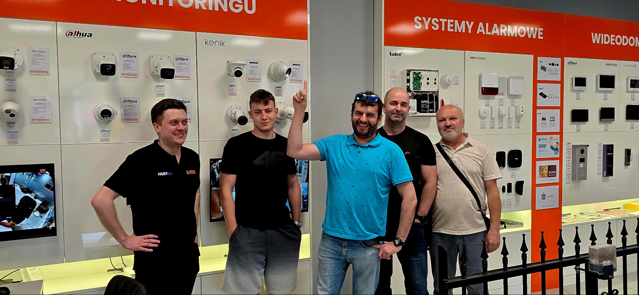 Ekspozycja systemów alarmowych, monitoringu i wideodomofonów z grupą mężczyzn. Widoczne kamery Dahua i Hikvision, alarmy Satel i wideodomofony Vidos X.