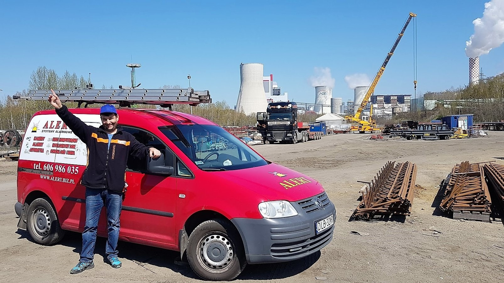 Uśmiechnięty monter w niebieskiej czapce pozuje obok czerwonego samochodu firmowego z logo 'ALERT SYSTEMY ALARMOWE' na tle przemysłowego krajobrazu z kominami elektrowni i dźwigiem.