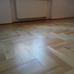 Ułożony parkiet w jodełkę francuską, jasne drewno, widoczny grzejnik na ścianie, listwy przypodłogowe.