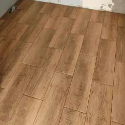 Ułożone brązowe płytki podłogowe imitujące drewno w rogu pomieszczenia z surowymi ścianami w trakcie remontu.