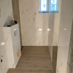 Wykończona łazienka z białymi marmurkowymi płytkami na ścianach i drewnopodobnymi panelami na podłodze, widoczne elementy instalacji sanitarnej i okno z zewnątrz.