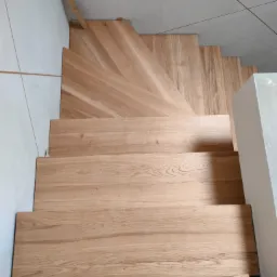 Drewniane schody z jasnego dębu, ułożone na betonowej podstawie, widok z góry, z fragmentem białej ściany z płytkami i surowym słupkiem balustrady.