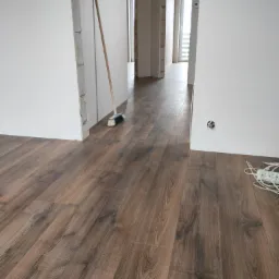 Wnętrze w trakcie prac wykończeniowych: ułożone panele podłogowe w odcieniach brązu, świeżo pomalowane białe ściany, widoczny fragment niezabezpieczonego gniazdka elektrycznego, oparta o ścianę...