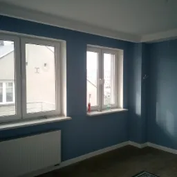 Pomalowane na niebiesko wnętrze pokoju z dwoma oknami, białymi ramami okiennymi i parapetami, grzejnikiem pod oknem, drewnianą podłogą i dekoracyjną listwą przypodłogową.