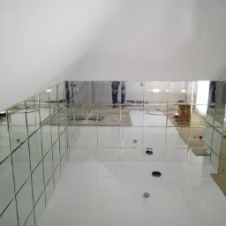 Montaż lustrzanych płytek w łazience na poddaszu, widoczne otwory na armaturę i odblask pracownika w lustrach.