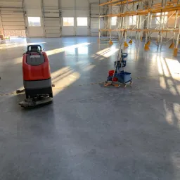 Czerwona maszyna do czyszczenia podłóg przemysłowych i wózek z mopem na tle hali magazynowej ze stalowymi regałami i wpadającym światłem słonecznym przez okna dachowe.