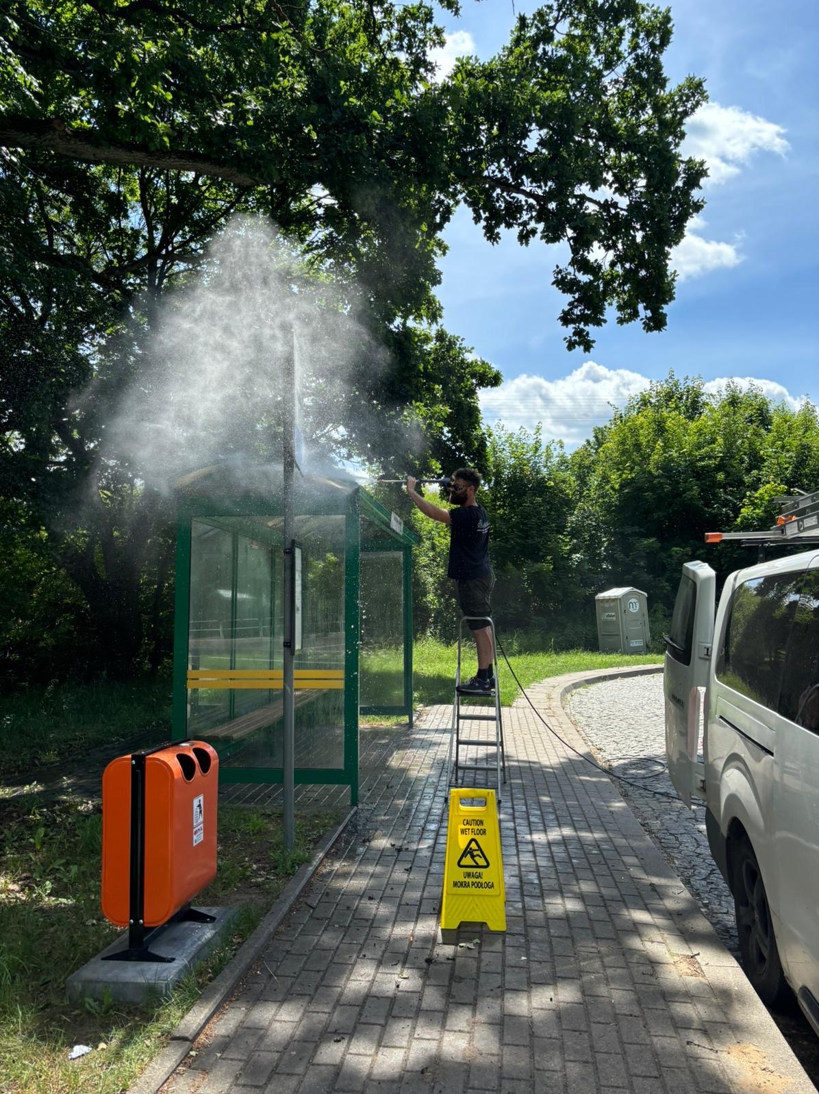 Pracownik na drabinie, w okularach przeciwsłonecznych, myje ciśnieniowo przystanek autobusowy z zieloną ramą, obok stoi pomarańczowy kosz na śmieci i znak ostrzegawczy 'Uwaga! Mokra podłoga', w tle...