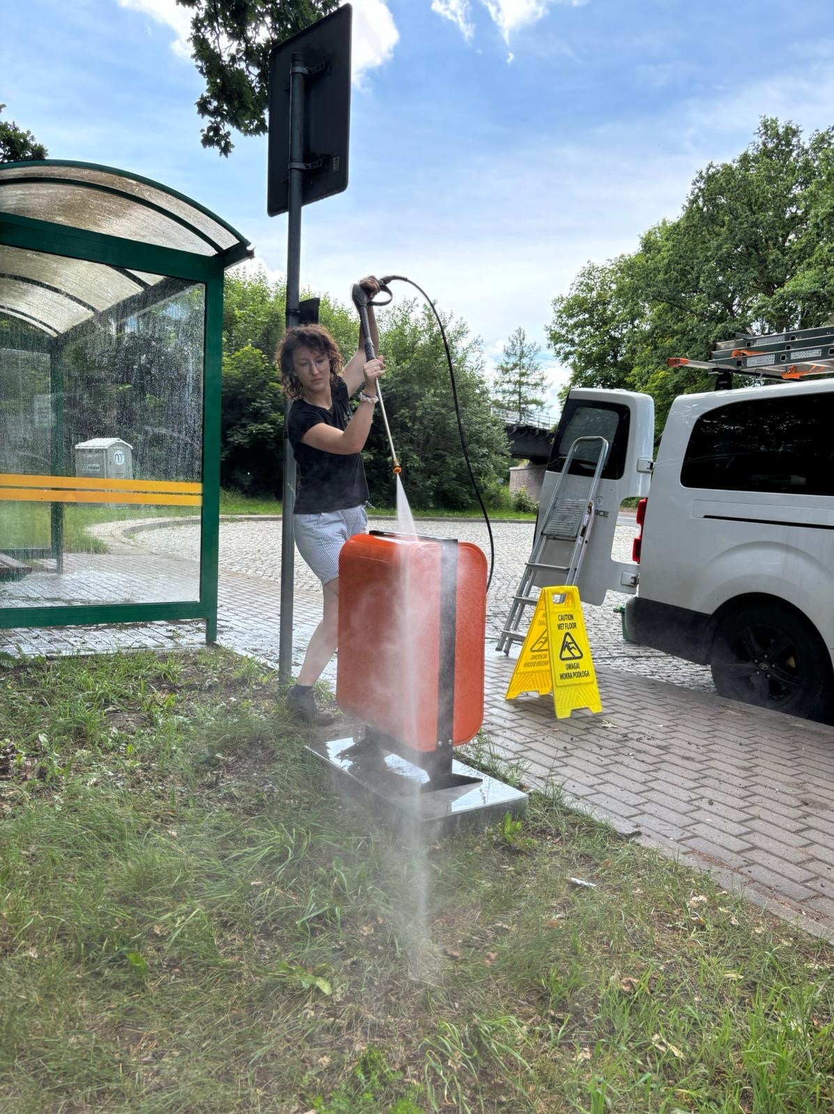 Kobieta czyści myjką ciśnieniową pomarańczowy pojemnik na śmieci na przystanku autobusowym, z widocznym znakiem ostrzegawczym o mokrej podłodze i samochodem dostawczym w tle.