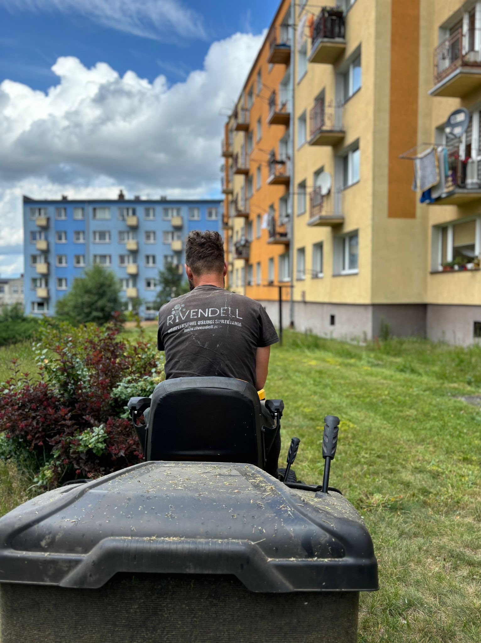 Mężczyzna siedzący na kosiarce traktorowej, z widocznym logiem firmy 'Rivendell' na koszulce, podczas koszenia trawnika przed blokami mieszkalnymi w słoneczny dzień.