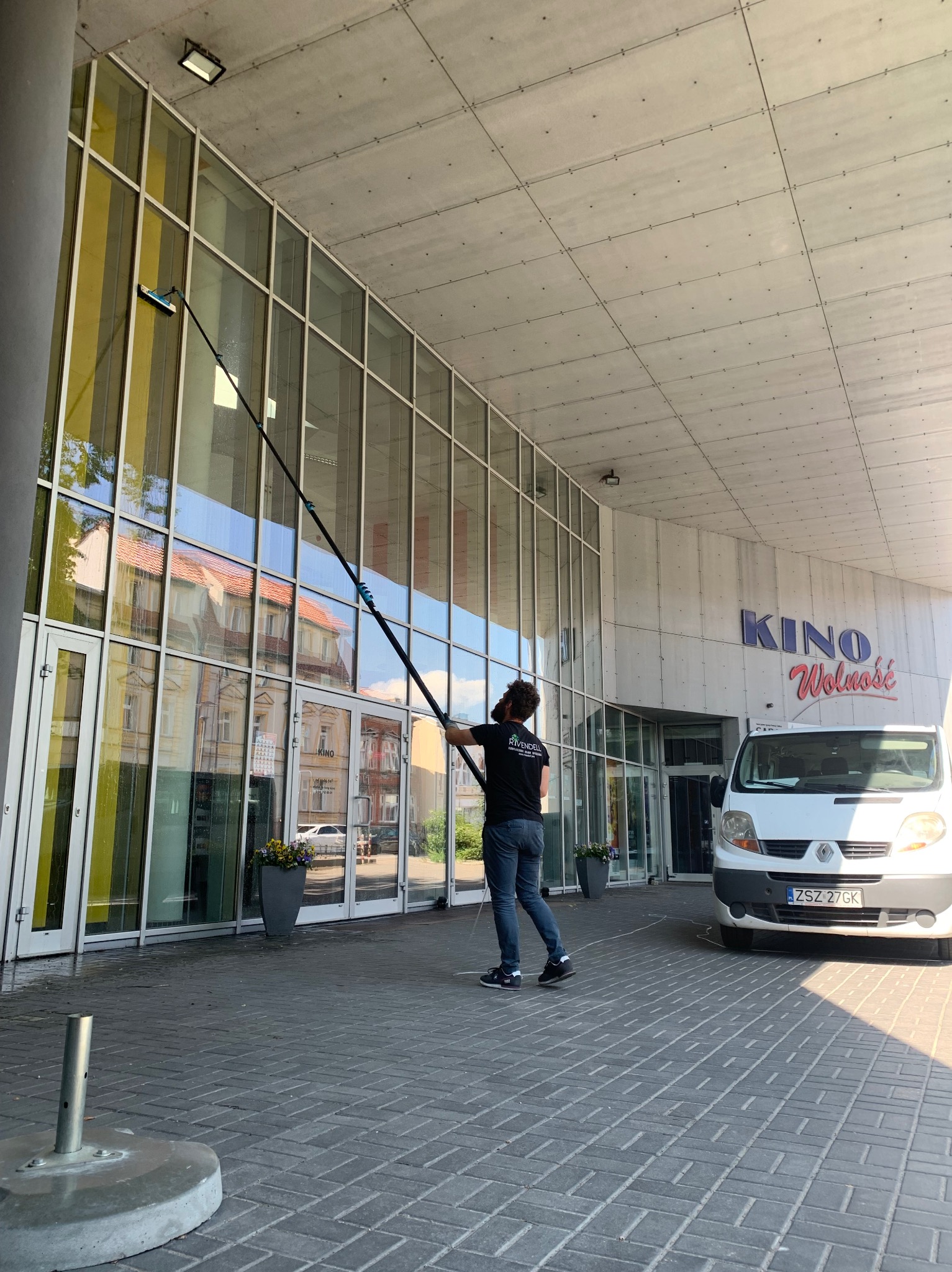 Pracownik w czarnej koszulce z logo firmy myje wysokie okna kina za pomocą długiego kija z myjką, na pierwszym planie zaparkowany biały bus.