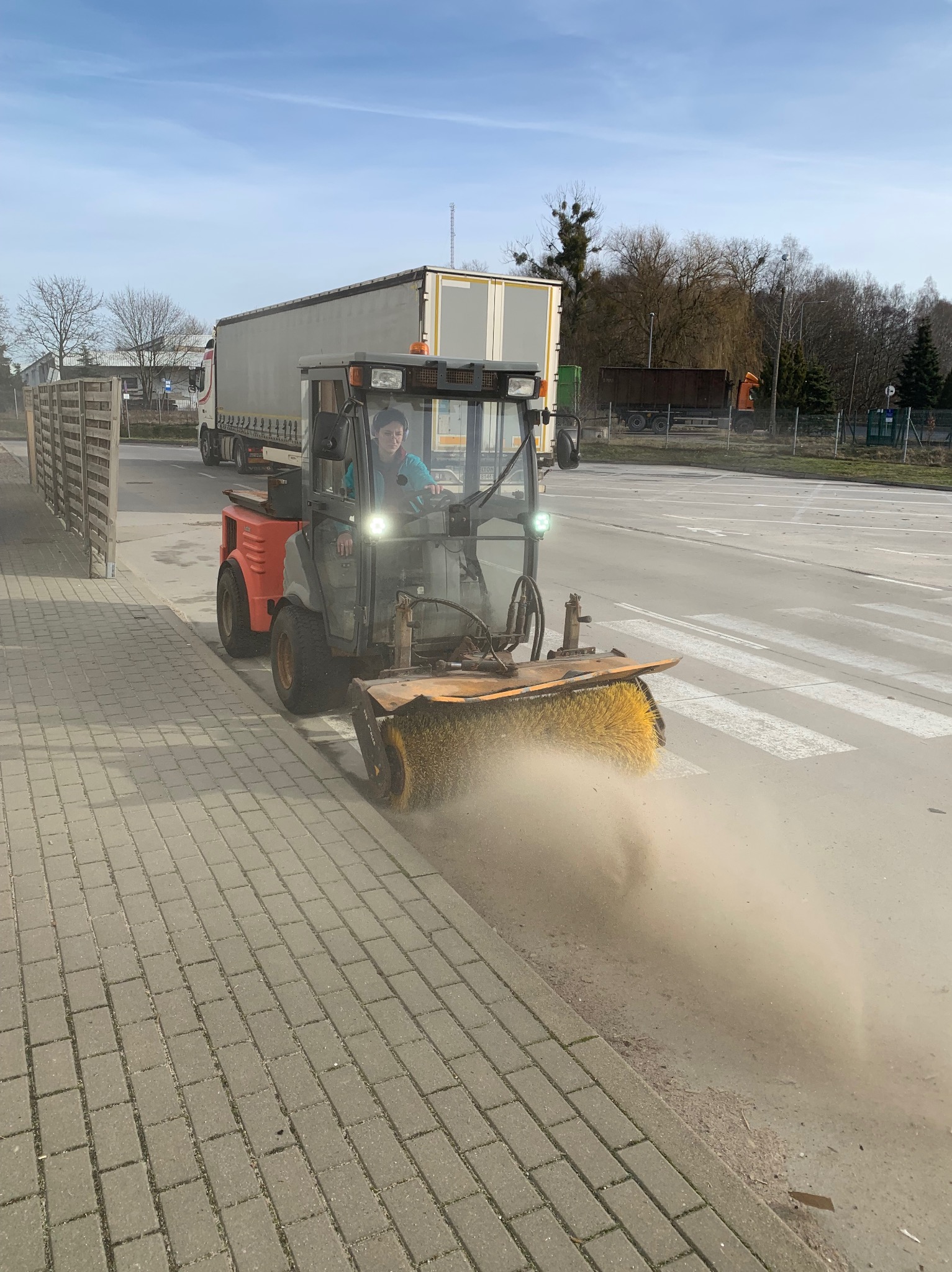 Operator w kabinie zamiatarki mechanicznej czyści krawężnik w Okonku, unoszący się pył, w tle zaparkowana ciężarówka.