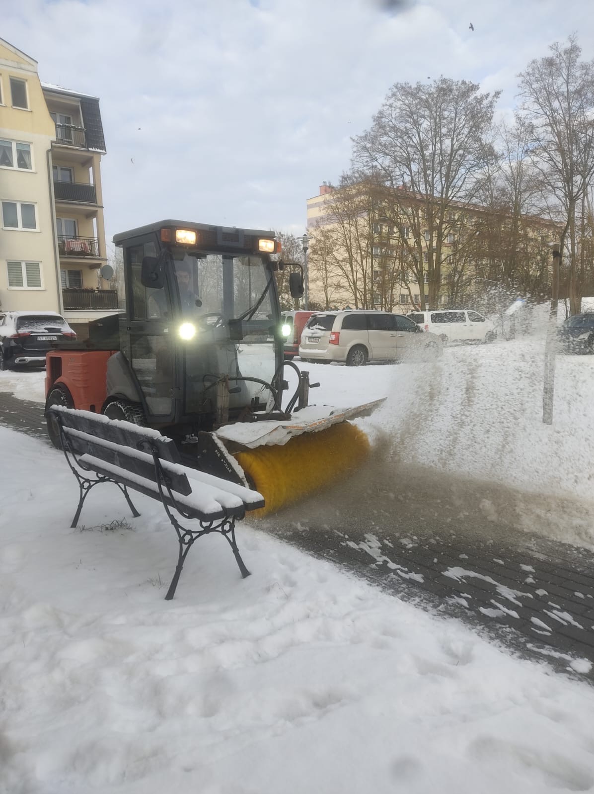 Pomarańczowo-szara zamiatarka wirnikowa usuwa śnieg z chodnika w pobliżu osiedla mieszkaniowego, obok zasypana śniegiem ławka parkowa.