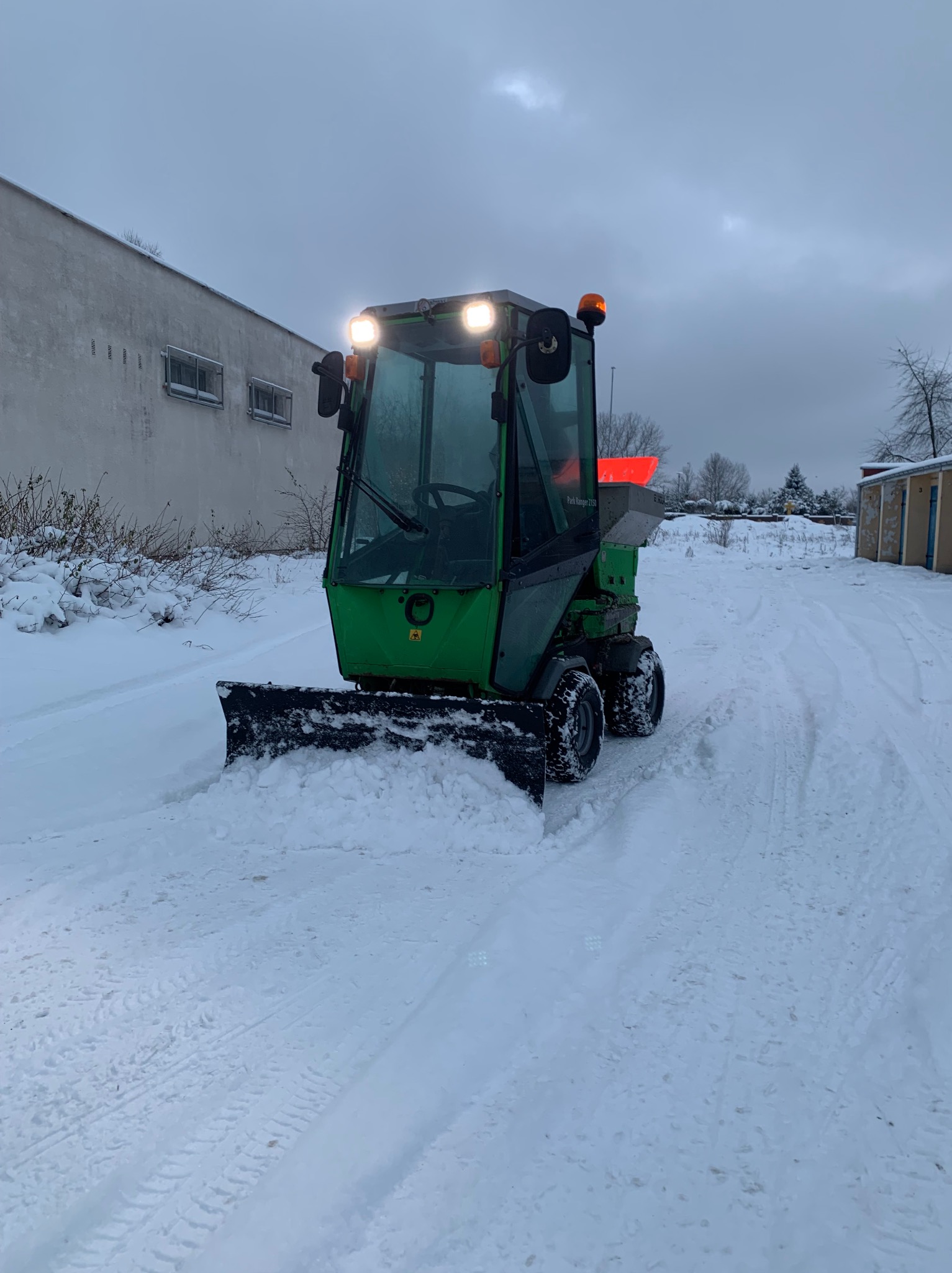 Zielony, kompaktowy traktor z pługiem odśnieżającym, usuwający śnieg z drogi w pochmurny dzień. Widoczne ślady opon na śniegu i budynki w tle.