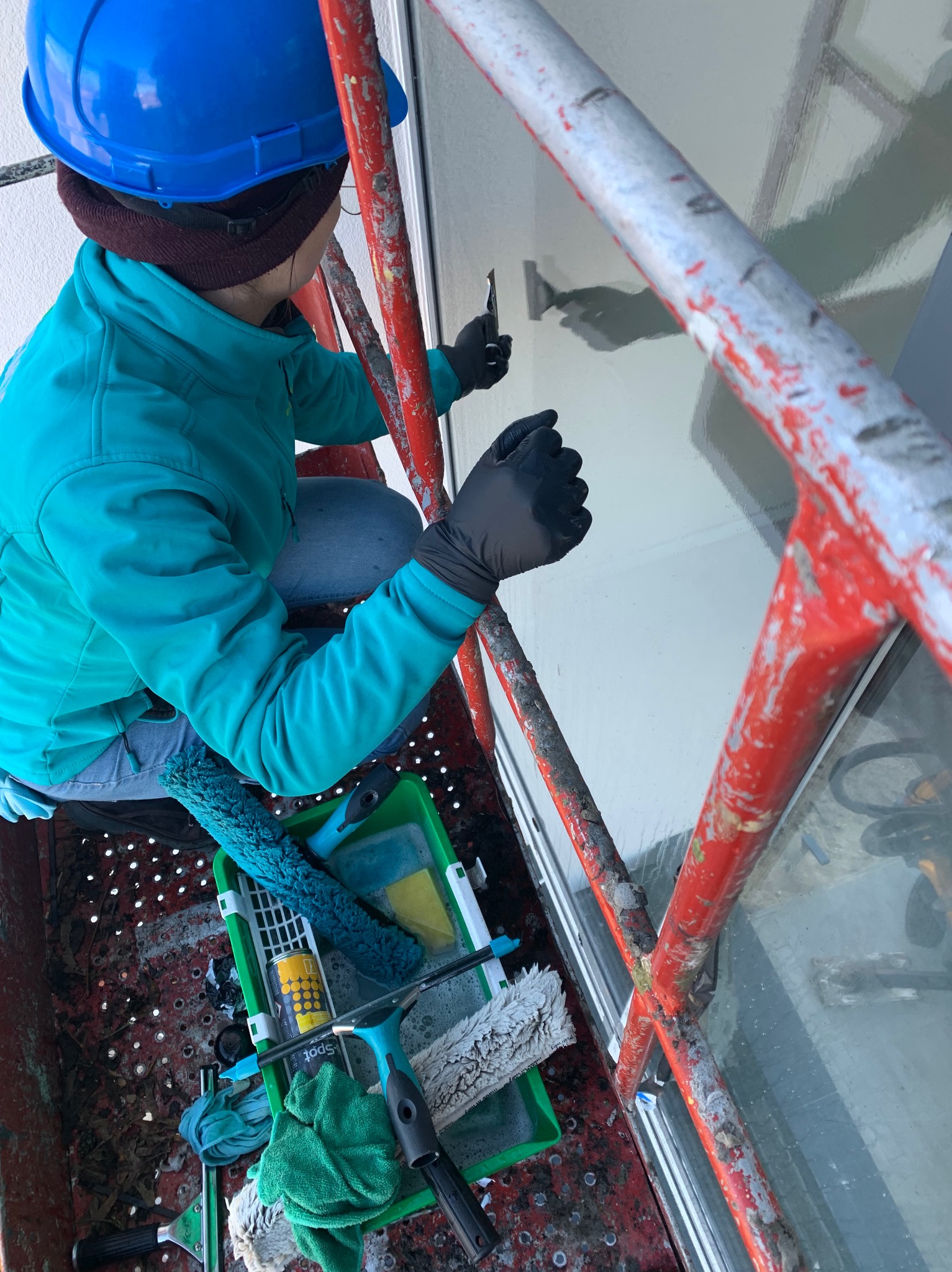 Pracownik w niebieskim kasku na rusztowaniu, czyści okno na wysokości, używając skrobaka; widoczne narzędzia w zielonym wiadrze i czerwone rusztowanie.