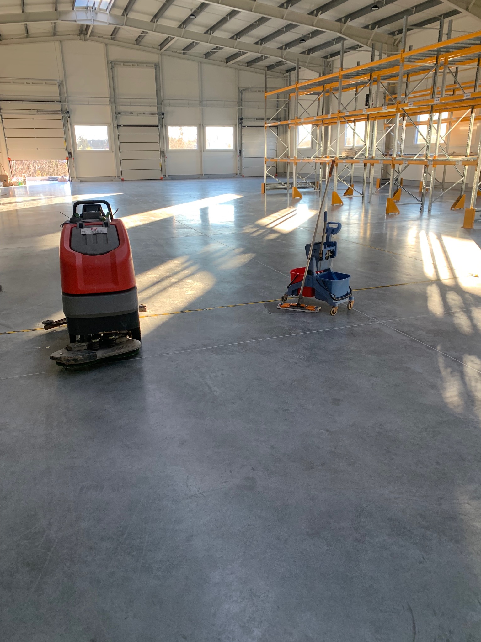 Czerwona maszyna do czyszczenia podłóg przemysłowych i wózek z mopem na tle hali magazynowej ze stalowymi regałami i wpadającym światłem słonecznym przez okna dachowe.