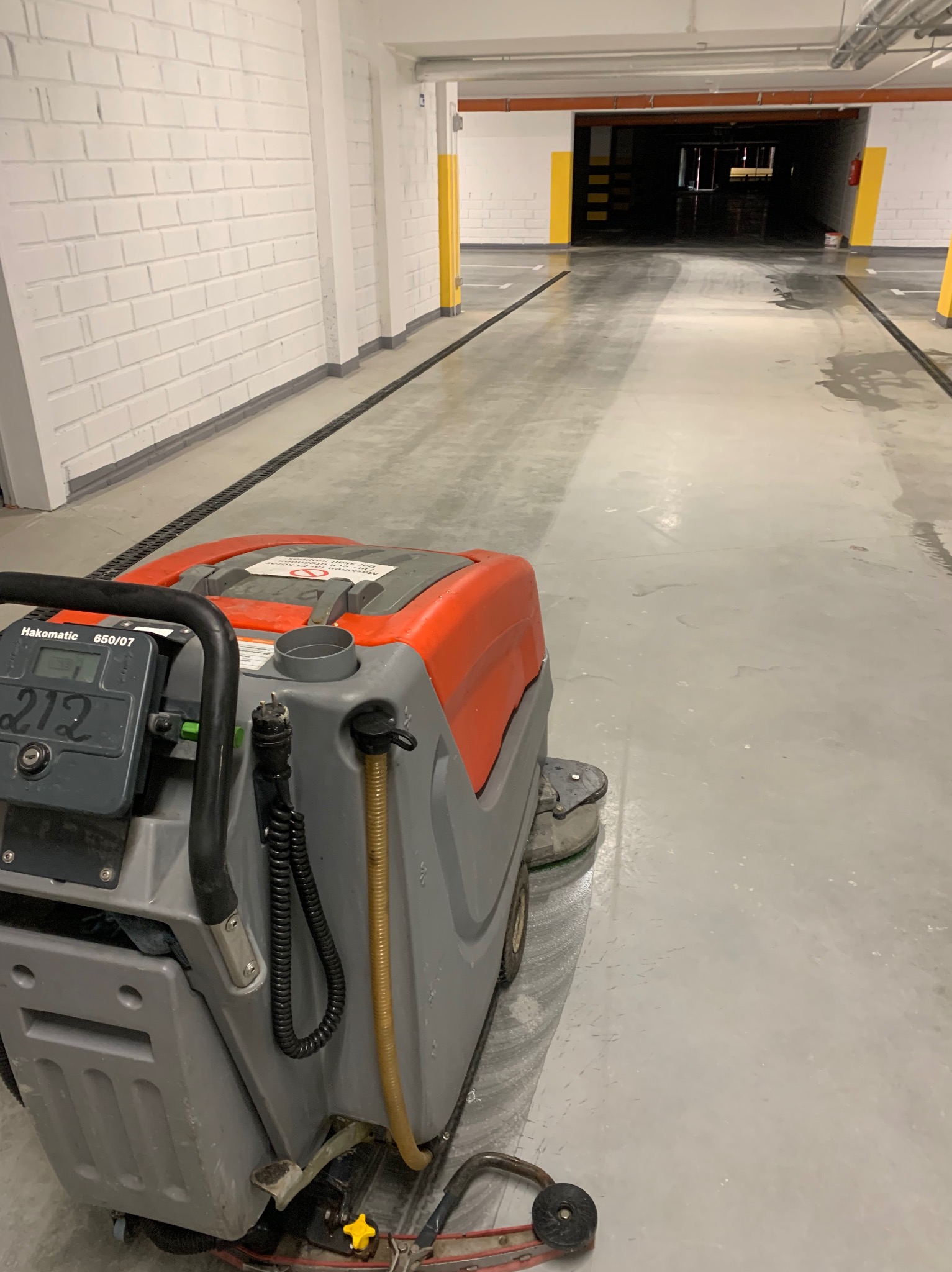 Szorowarka Hakomatic 650/07 z widocznym wyświetlaczem 212, czyszcząca posadzkę w garażu podziemnym z białymi ścianami z cegły i żółtymi słupami.