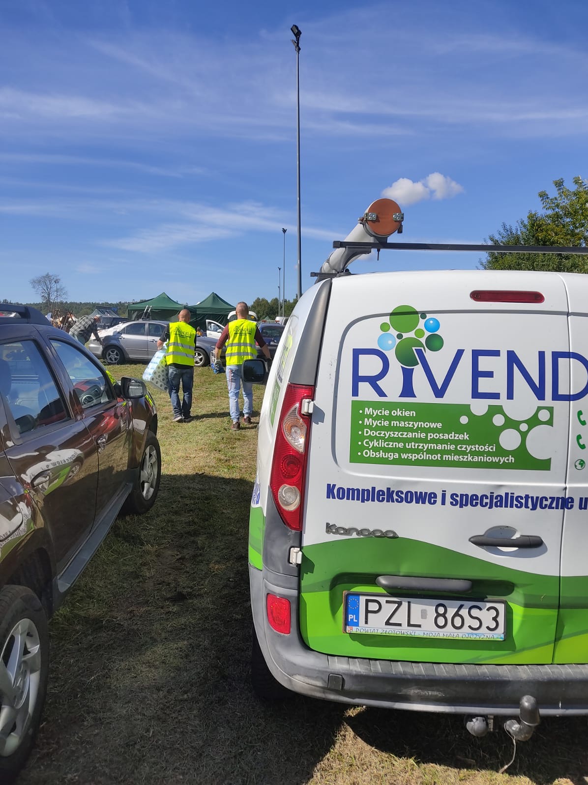 Furgonetka firmy Rivend z informacjami o myciu okien i doczyszczaniu posadzek, z pracownikami w żółtych kamizelkach niosącymi rolki papieru w tle, na trawiastym terenie.