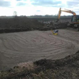 Żółta koparka JCB przygotowuje teren pod budowę, ubijarki wibracyjne zagęszczają ziemię w wykopie, widoczne ślady po ubijaniu, na horyzoncie pole i drzewa pod pochmurnym niebem.