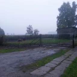 Prosta, metalowa brama wjazdowa z furtką w Strzelinku, widoczna z perspektywy podjazdu w pochmurny dzień.