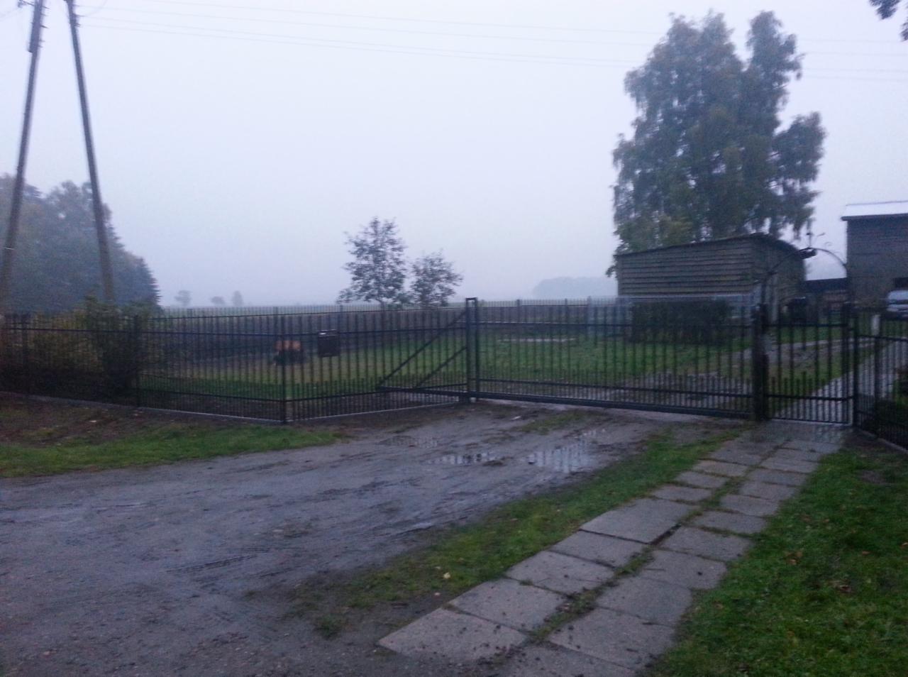 Prosta, metalowa brama wjazdowa z furtką w Strzelinku, widoczna z perspektywy podjazdu w pochmurny dzień.