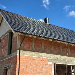 Ciesielstwo-Dekarstwo SŁAW-DACH - Dom w trakcie budowy z widocznym dachem pokrytym czarną dachówką i zamontowanymi rynnami. Murowana konstrukcja z czerwonej cegły i betonu pod błękitnym niebem.