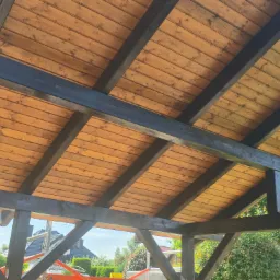 Drewniana konstrukcja z belek i desek, pomalowana na brązowo i czarno, widoczna od spodu, z fragmentem krajobrazu w tle. Widoczny fragment drabiny.