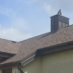 Brązowy dach z gontów bitumicznych i ciemne rynny, z kominem w nowoczesnym stylu na tle błękitnego nieba. Widok z dołu, akcentujący detale wykończenia.