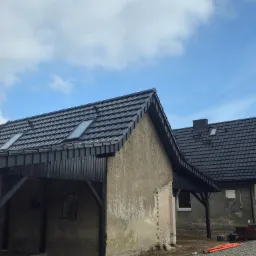 Dwa budynki z nowym, czarnym dachem z dachówką i oknami dachowymi. Jeden z budynków posiada drewnianą konstrukcję wspartą na słupach. Błękitne niebo z chmurami w tle.