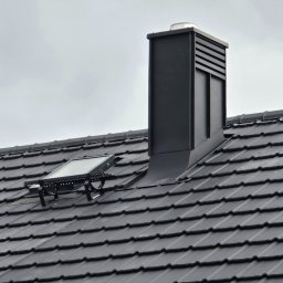 Ciesielstwo-Dekarstwo SŁAW-DACH - Grafitowy dach z dachówki ceramicznej z oknem połaciowym i nowoczesnym kominem wentylacyjnym na tle pochmurnego nieba. Widok z dołu.