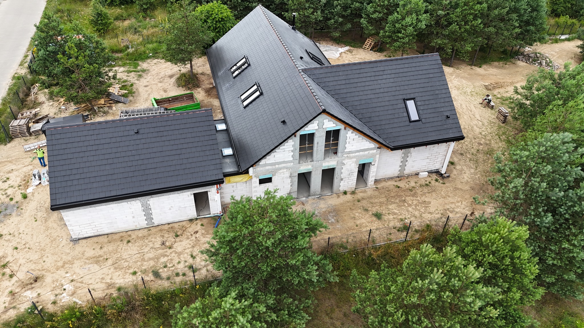 Widok z lotu ptaka na budynek w trakcie budowy z zamontowanym, ciemnoszarym pokryciem dachu z dachówki ceramicznej, widoczne okna dachowe i niezakończone ściany z białego bloczka.